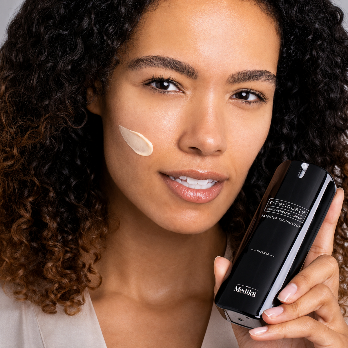 Жінка тримає крем Medik8 r-Retinoate Intense із текстурою засобу на щоці