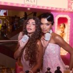 Дві жінки тримають флакони KAYALI Boujee Kitty Caramel Milk на рожевому парфумерному стенді бренду