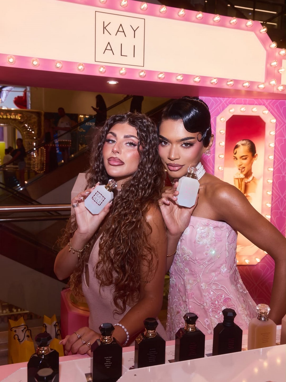 Дві жінки тримають флакони KAYALI Boujee Kitty Caramel Milk на рожевому парфумерному стенді бренду